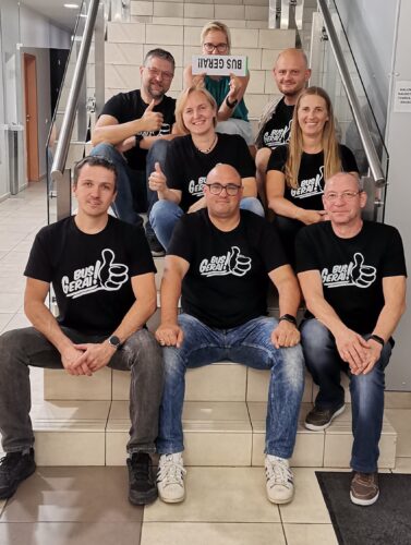 8 žmonės susėdę ant laiptų, su vienodais juodais marškinėliais su užrašu "Bus gerai!". Auksinio proto žaidimai