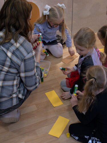 Vaikai ir bibliotekos darbuotoja klijuoja spalvotus lapelius prie sienos.