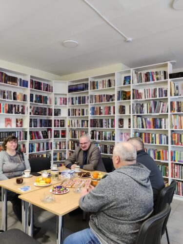 susitikimas bibliotekoje. Prie stalo sėdi žmonės, vaišinasi kava ar arbata. Stalas nukrautas įvairiomis vaišėmis.