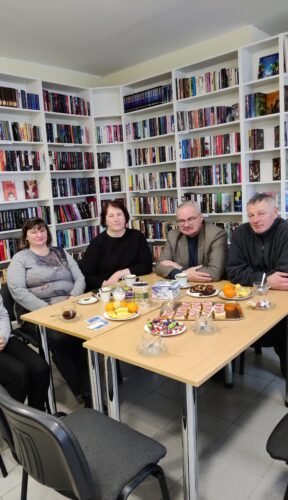 susitikimas bibliotekoje. Prie stalo sėdi žmonės, vaišinasi kava ar arbata. Stalas nukrautas įvairiomis vaišėmis.