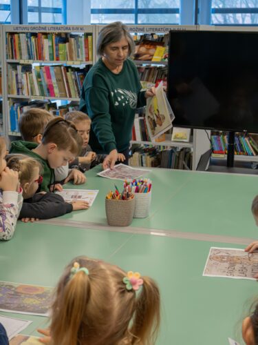 Edukacija bibliotekoje. Vaikai sėdi prie stalo, bibliotekos darbuotoja dalina lapus su atspausdintais piešiniais.