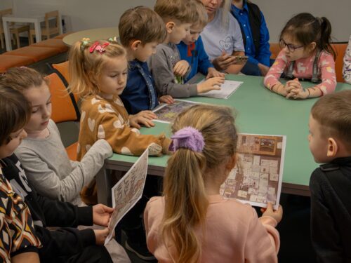 Vaikai sėdi prie stalo, apžiūrinėja atspausdintus piešinius, aptarinėja tai ką mato.