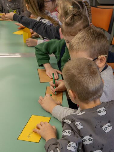 Vaikai sėdi prie stalo, piešia, rašo ant spalvotų lapukų.