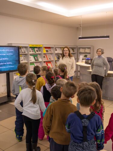 Vaikai stovi bibliotekos fojė. Dvi bibliotekos darbuotojos stovi prieš vaikus ir su jais šneka.