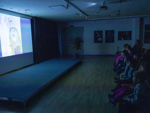 Vaikai sėdi auditorijoje ir žiūri per projektorių rodomą filmą