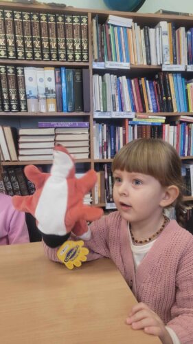 Mergaitė bibliotekoje sėdi prie stalo, žaidžia su pliušiniais žaislais.