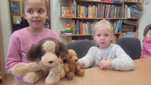 Mergaitės bibliotekoje sėdi prie stalo, žaidžia su pliušiniais žaislais.