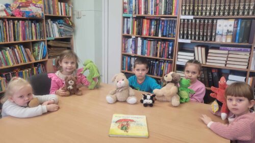 Vaikai bibliotekoje sėdi prie stalo, žaidžia su pliušiniais žaislais.